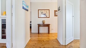 Un pasillo con piso de madera, paredes blancas y una puerta a la derecha. Hay una pequeña mesa con un florero y un cuadro en la pared.