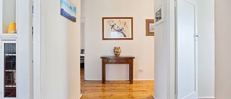 A hallway with wooden flooring, white walls, and a door on the right. There is a small table with a vase and a framed picture on the wall.
