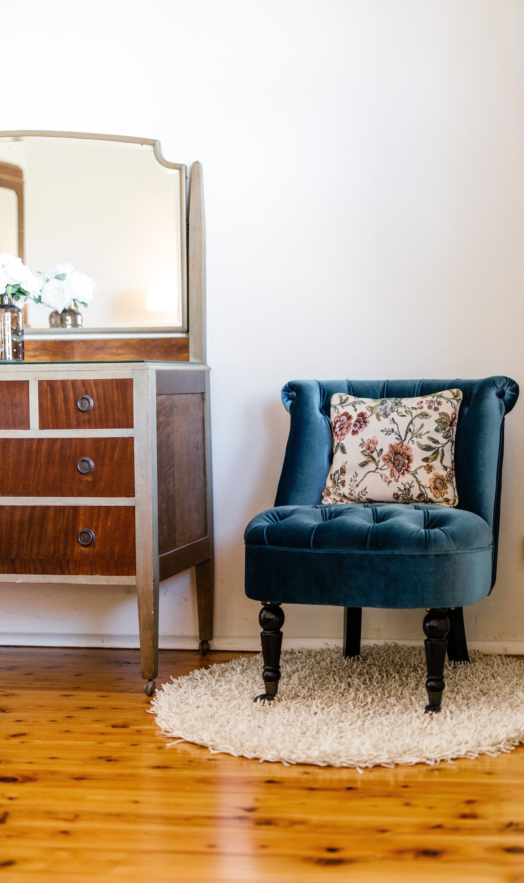 A room with a wooden dresser, a blue upholstered chair with a floral pillow, and a white shag rug on a wooden floor.