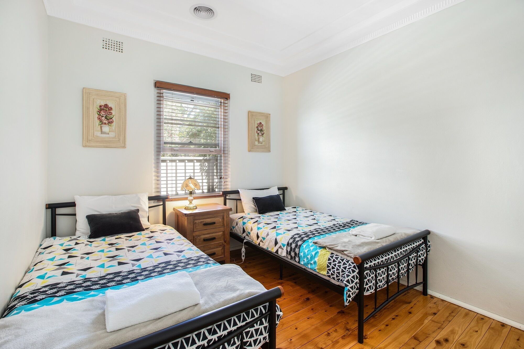 Une chambre avec deux lits, une table de chevet, une fenêtre avec des stores et deux tableaux encadrés accrochés au mur.