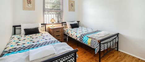 A bedroom with two beds, a nightstand, a window with blinds, and two framed pictures on the wall.
