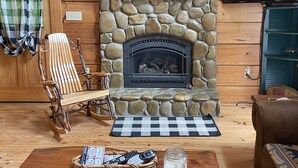 Un salon chaleureux avec une cheminée en pierre, des murs en bois, un tapis à carreaux, un fauteuil à bascule et un téléviseur.