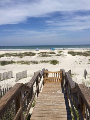A wooden boardwalk leading to a beach with sand dunes and a clear sky.