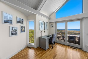 Una habitación con escritorio, silla y ventanas amplias que dan a una terraza con mobiliario exterior.