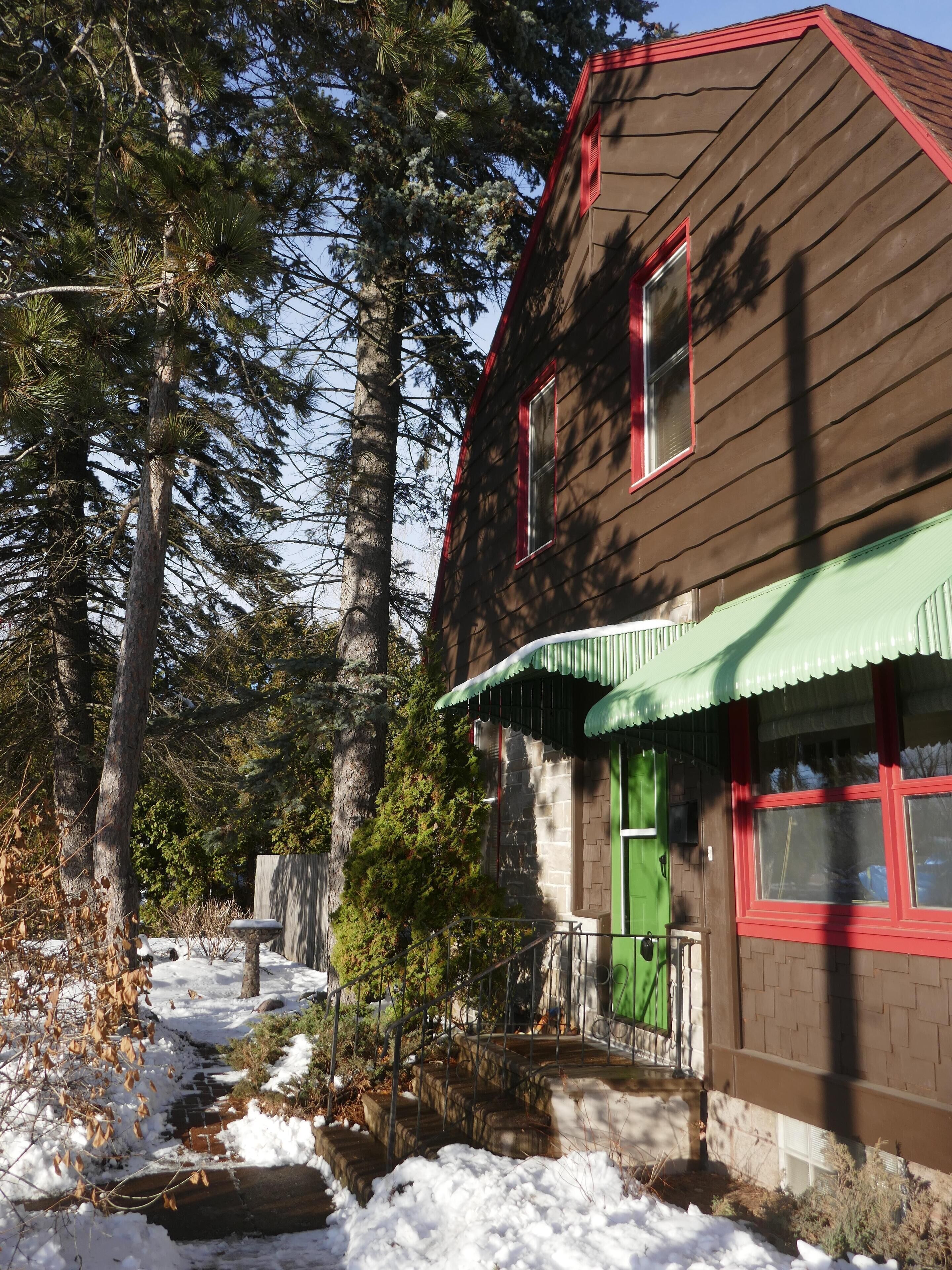 Une maison en bois avec une porte verte et des bordures rouges, entourée d’arbres et bordée d’un chemin enneigé.