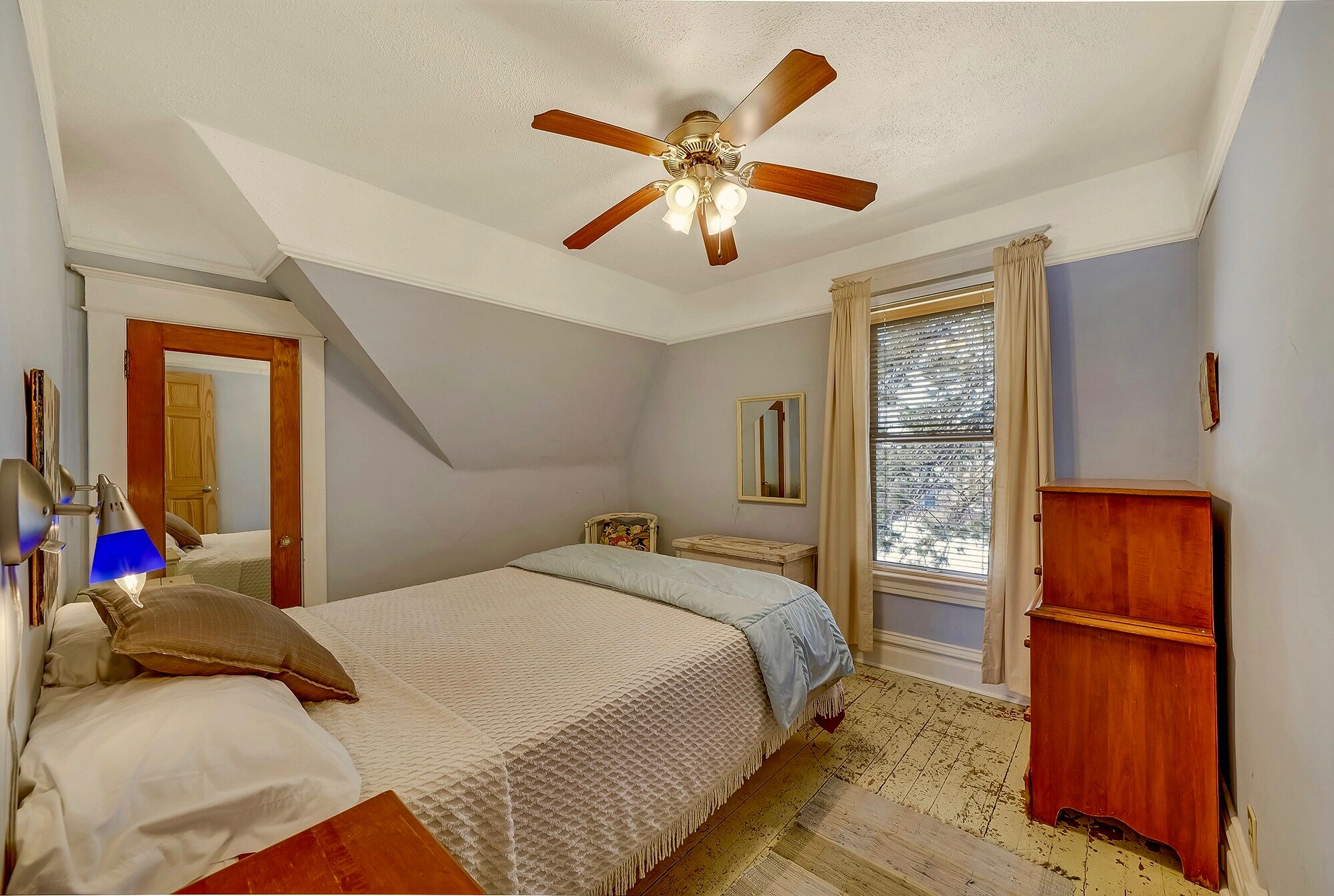 A bedroom with a bed, a ceiling fan, a window with blinds, a wooden dresser, and a desk with a lamp.