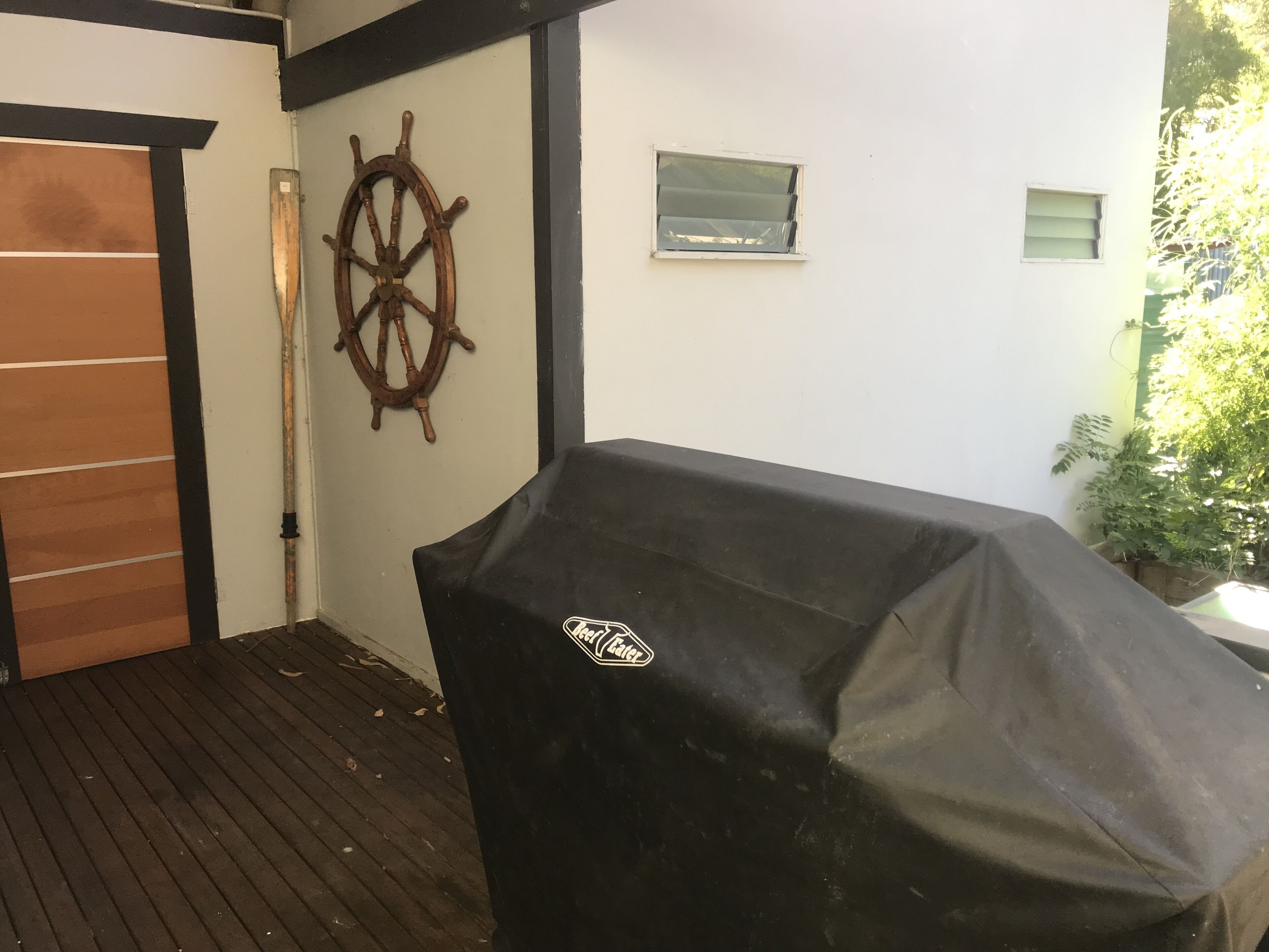 A covered barbecue grill, a wooden ship's wheel, and a small window on a white wall.