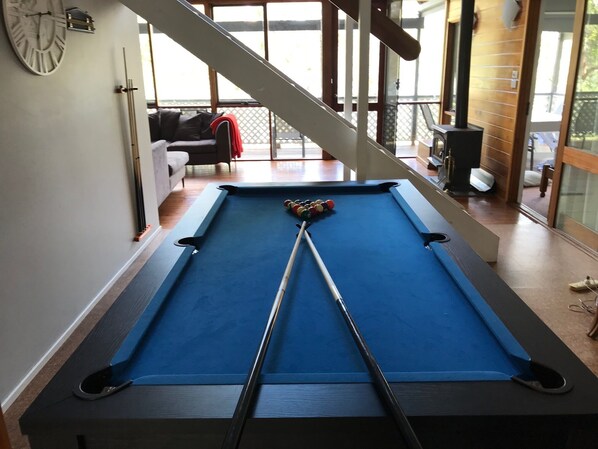 A pool table in a room with a staircase, a clock on the wall, and a sofa in the background.