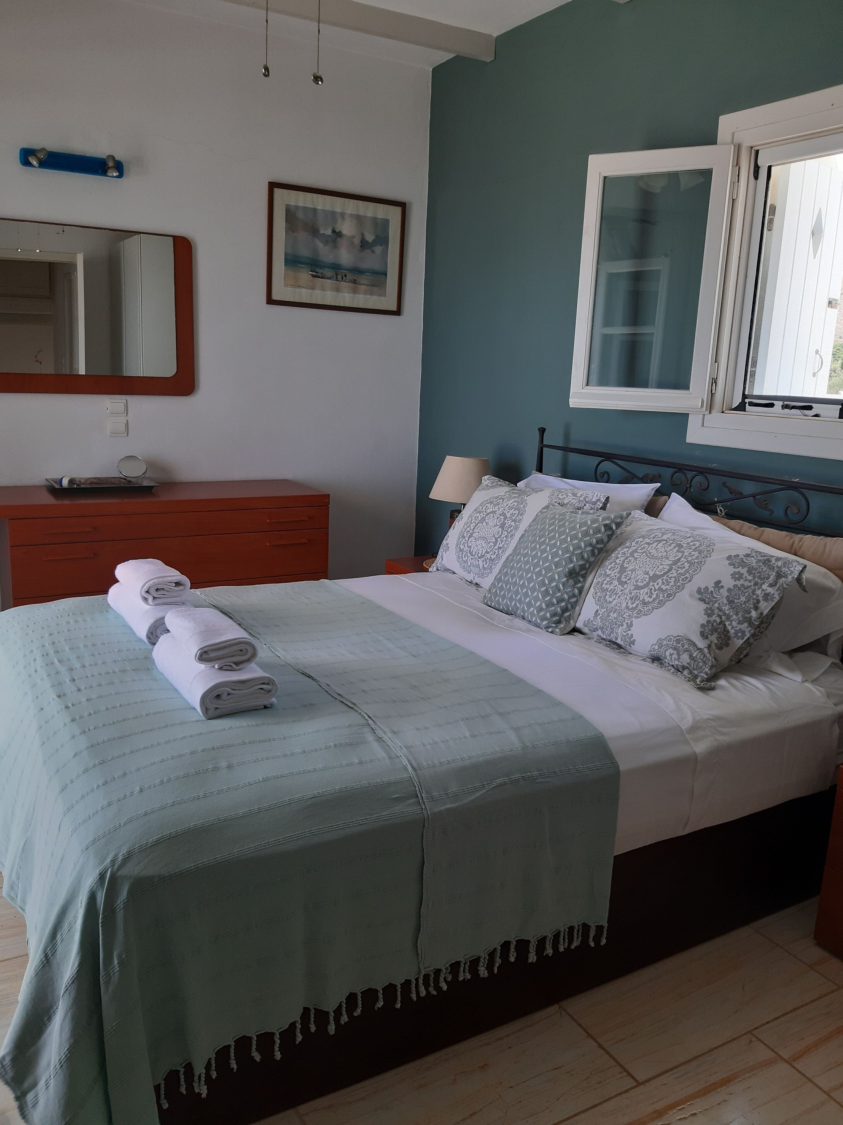 Een netjes opgemaakt bed met een blauw sprei en kussens, een houten dressoir, een spiegel en een raam met uitzicht op de omgeving.
