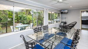 A spacious room with a large glass table surrounded by chairs, large windows offering a view of greenery, and a ceiling fan. - Beach Hound (pets welcome) (Toogoom)