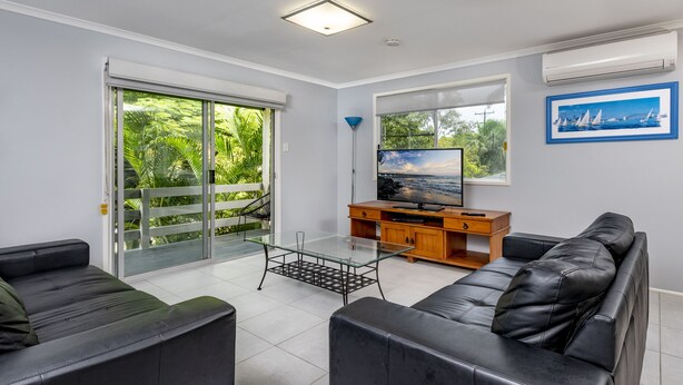 A modern living room with a large TV, leather sofas, a glass coffee table, and a balcony with a view of greenery. - Beach Hound (pets welcome) (Toogoom)