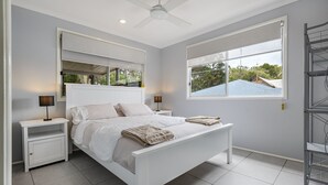 A modern bedroom with a white bed, two bedside tables, a ceiling fan, and a large window with a view of the outside. - Beach Hound (pets welcome) (Toogoom)