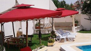 Un área junto a la piscina con sombrillas rojas, sillones blancos y una pequeña mesa con sillas.
