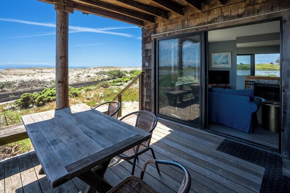 A wooden deck with a table and chairs, a sliding glass door leading to a room with a bed and a TV, and a view of the landscape.
