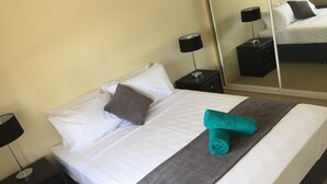 A neatly made bed with white linens, a gray throw blanket, and a teal towel. There are two black nightstands with lamps, one on each side of the bed. - Golf Course Apartment/Within Trinity Links Resort/ Selfcontained (White Rock)