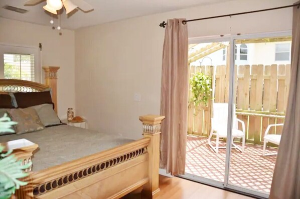 Une chambre avec un lit, un ventilateur de plafond, une fenêtre avec des stores et une porte-fenêtre coulissante donnant sur une terrasse extérieure équipée de chaises et entourée d’une clôture en bois.