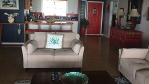 Een woonkamer met een beige bank, een salontafel met afstandsbedieningen, en een rode deur op de achtergrond.