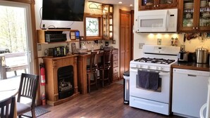 Una cocina compacta con estufa a gas, microondas y un refrigerador pequeño. Hay una mesa de comedor de madera con sillas, una chimenea y un televisor montado en la pared.