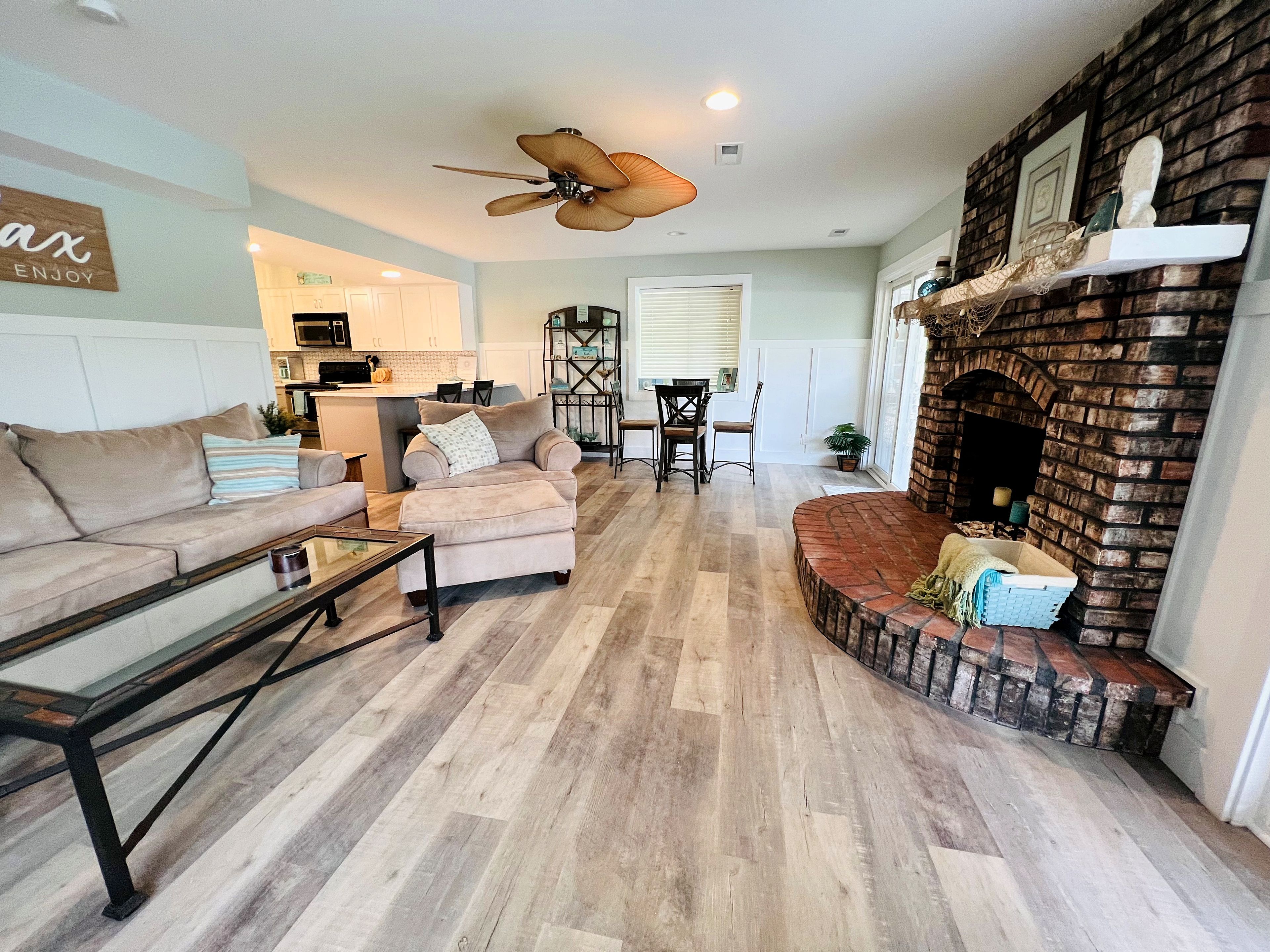 A living room with a fireplace, a ceiling fan, and a kitchen in the background.