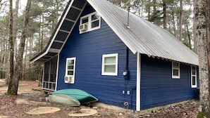 A small, single-story house with a steeply pitched roof, surrounded by a forest.