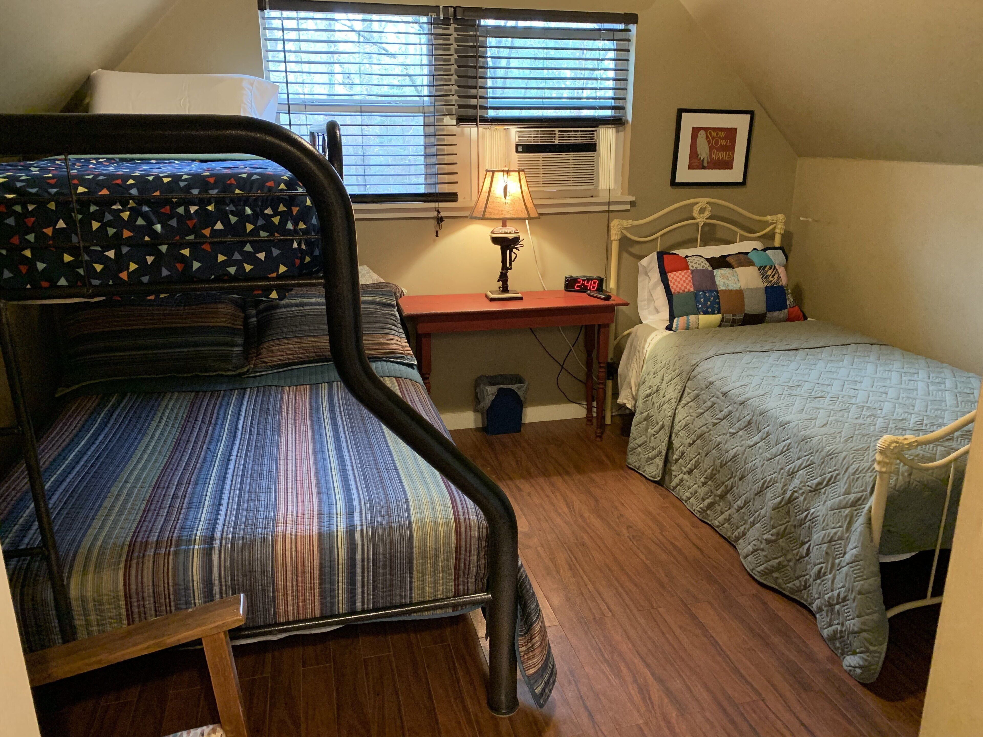 A dormitory room with a bunk bed, a desk with a lamp, a clock, and a window with blinds.