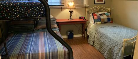 A dormitory room with a bunk bed, a desk with a lamp, a clock, and a window with blinds.