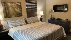 A neatly made bed with striped bedding, a bedside table with a lamp, a TV mounted on the wall, and a window with blinds.
