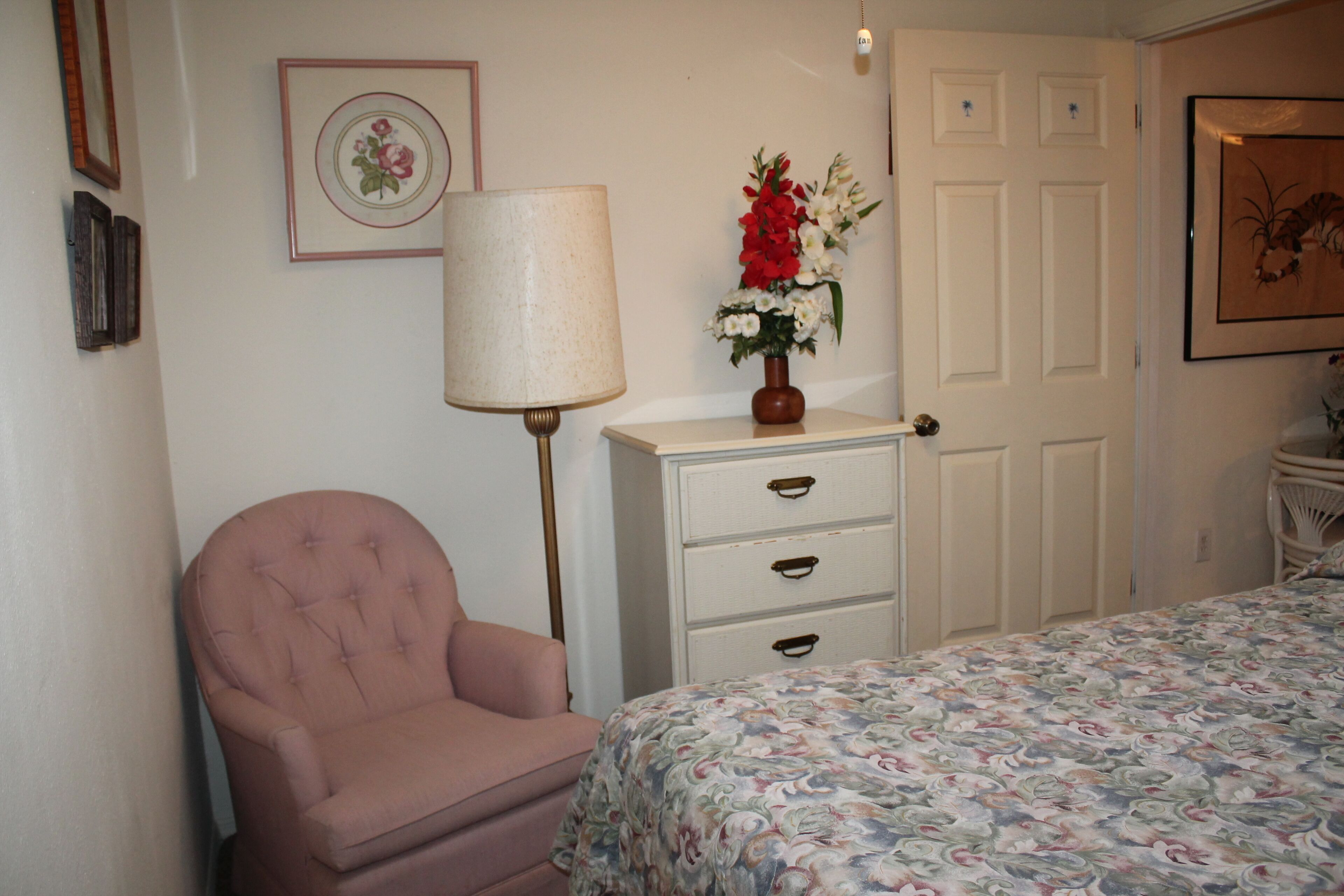 Un dormitorio con un sillón rosa, un aparador con cajones, una lámpara y una colcha estampada.