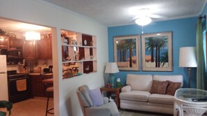 A living room with a sofa, armchair, and a glass coffee table. There is a kitchen area with cabinets and a microwave. Two framed pictures of palm trees are on the wall. -  Country  Cottage Home (Bradenton)