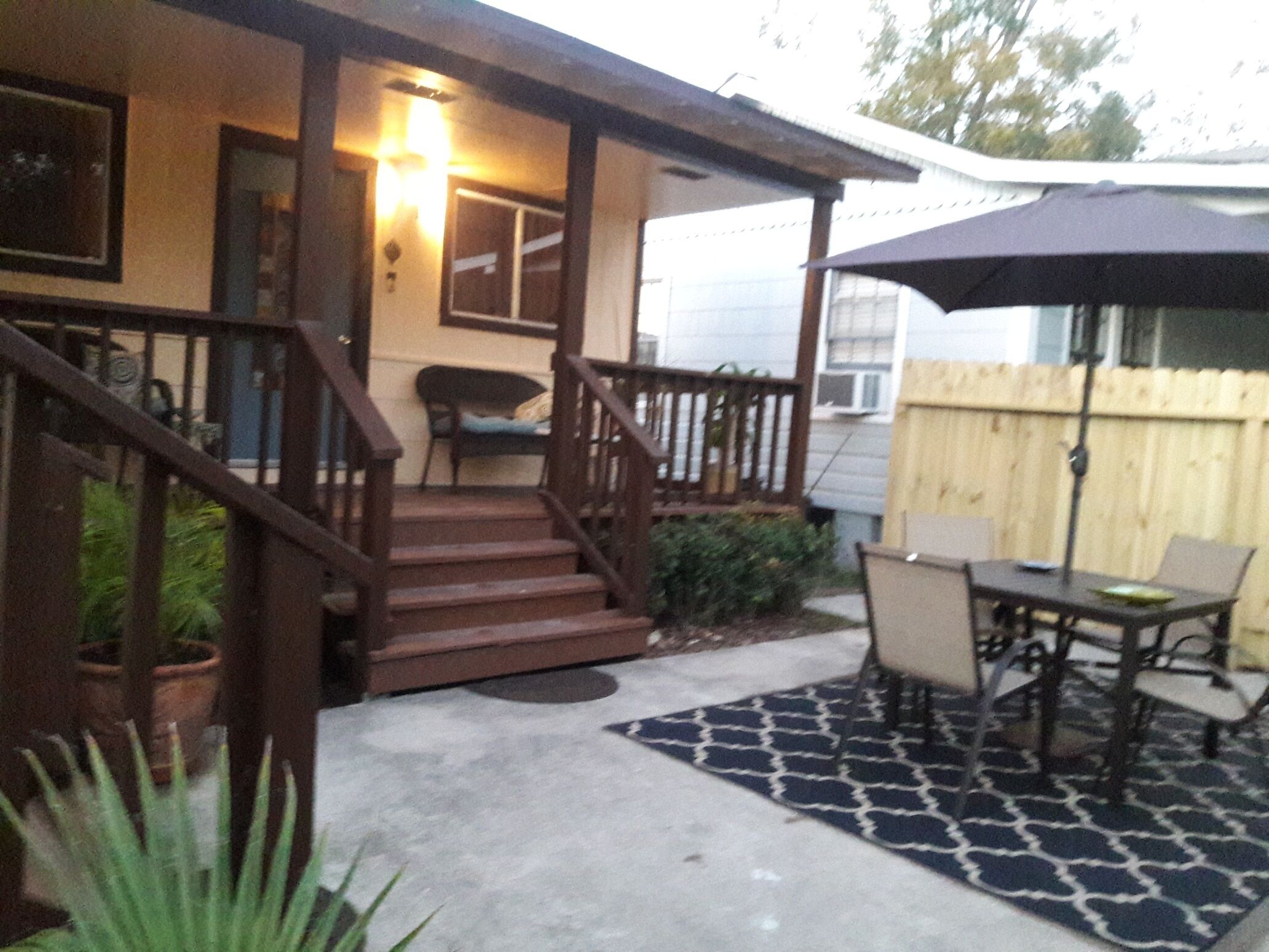 A covered patio area with a wooden staircase leading to a porch, a dining table with chairs, and a large umbrella.