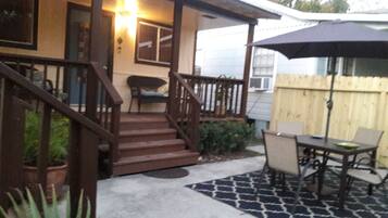 A covered patio area with a wooden staircase leading to a porch, a dining table with chairs, and a large umbrella.