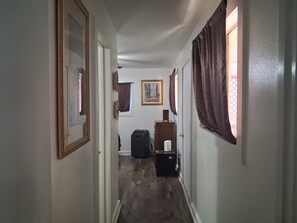 A narrow hallway with wooden flooring, a door on the left, a window with curtains on the right, and a framed picture on the wall.