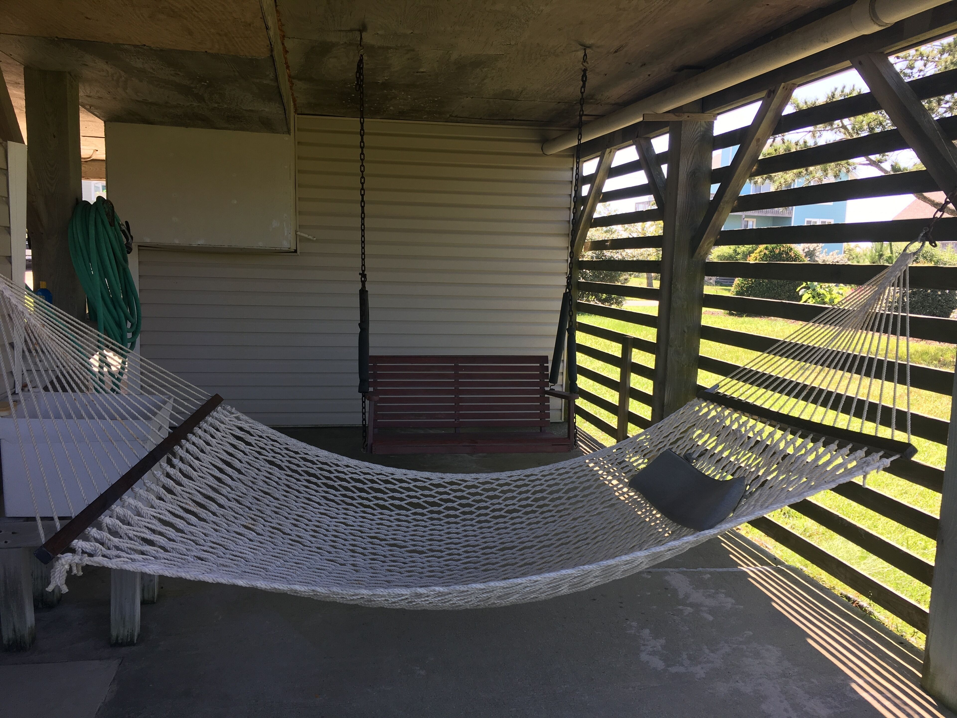 Riippumatto puisen pergola-rakenteen alla, jossa on myös penkki ja taustalla näkyy vihreä puutarhaletku.
