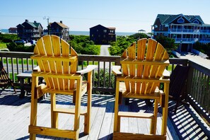 Two wooden Adirondack chairs on a wooden deck overlooking a coastal area with houses and a view of the ocean.