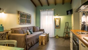 A cozy living area with a sofa, wooden stools, a dining table, and a kitchen with a stove and cabinets. - Charming and traditional small stone house. (Corfu)