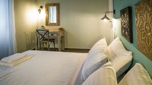 A neatly made bed with white linens, a desk with a chair, a wall-mounted lamp, and a decorative wall piece. - Charming and traditional small stone house. (Corfu)