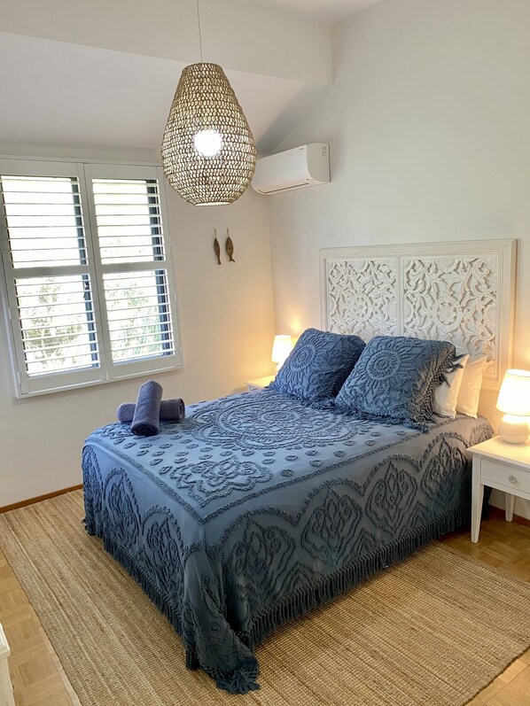 A bedroom with a bed, a hanging light fixture, a window with blinds, and a wall-mounted air conditioner.