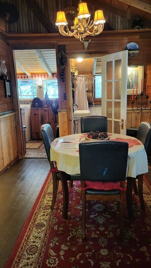 Interior de madera con una mesa de comedor lista para cuatro personas, rodeada de sillas. Un candelabro cuelga del techo, y una decoración rústica incluye un cráneo de oso y un aparador de madera.