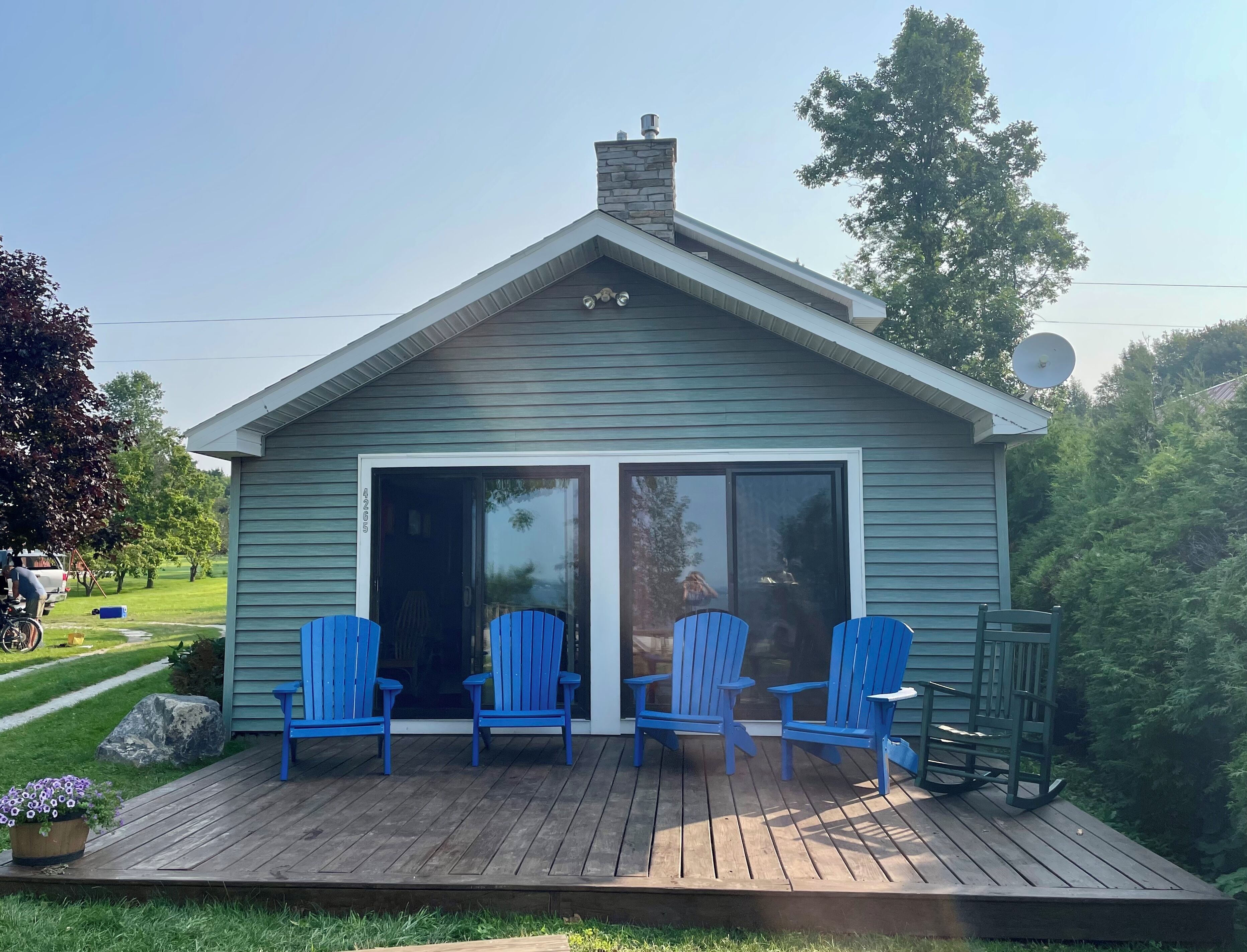 Una cabaña pequeña de azul claro con un muelle de madera, dos sillas Adirondack azules y una antena parabólica.