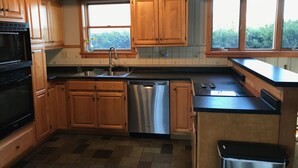 A kitchen with wooden cabinets, a stainless steel dishwasher, a black microwave, and a window with a view of greenery. - Slow down…You’re on Island Time (Isle La Motte)