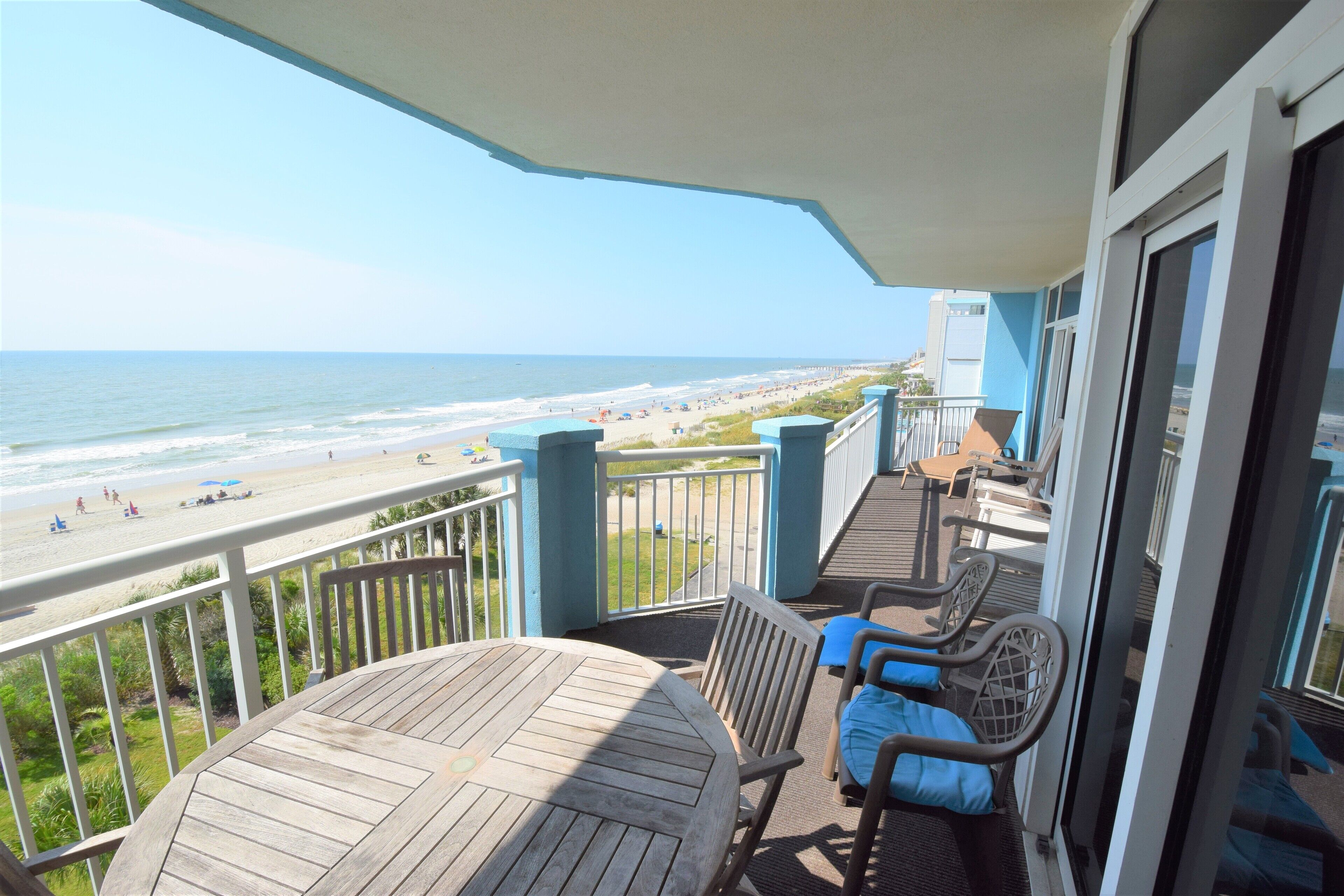 A balcony with a view of the beach, a table, and two chairs.