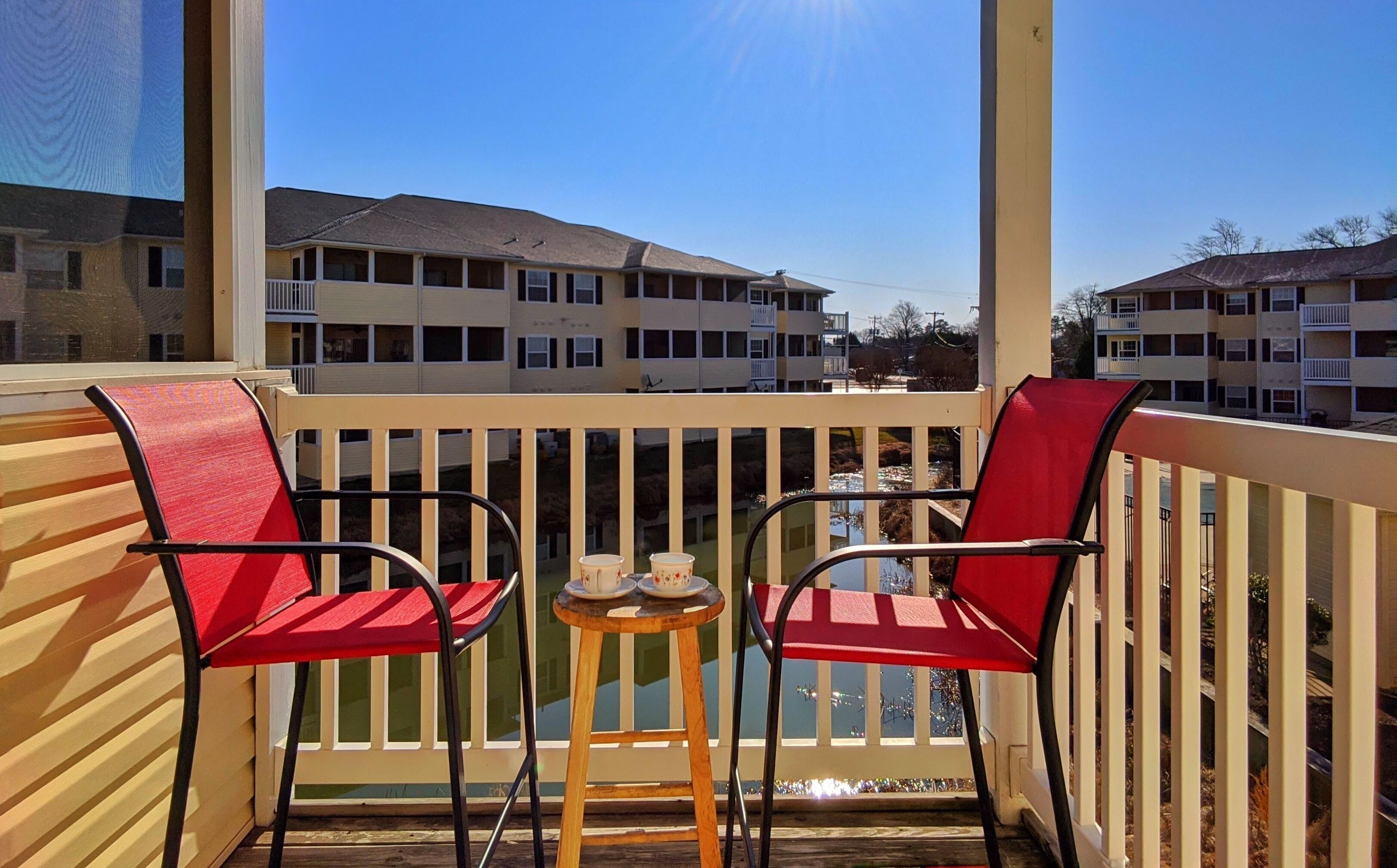 A balcony with two red chairs and a small table with cups, overlooking a row of multi-story buildings.