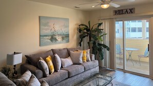 A living room with a grey sofa, a painting, a large plant, a ceiling fan, and a sliding glass door leading to an outdoor area. - Spacious Condo with Pool Near Beach- New Floors and Furniture!  (Rehoboth)