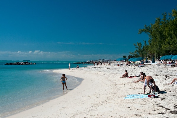 A beach with white sand, clear turquoise water, and a few people. There are umbrellas and beach chairs on the sand.