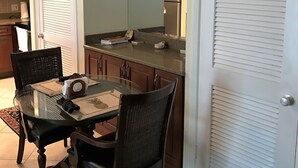 Comedor con mesa de vidrio, sillas y un reloj. Zona de cocina con armarios y refrigerador.