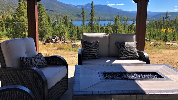 A covered outdoor seating area with two cushioned chairs and a fire pit, overlooking a mountainous landscape and a lake.