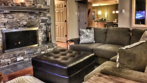 A living room with a stone fireplace, a sectional sofa, and a black leather ottoman. - Perfect for Large Groups • Hot Tub • Fire Pit • Fast Wi-Fi & Sparkling Clean (Pisgah Forest)