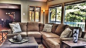 A spacious living room with a large L-shaped sectional sofa, a matching ottoman, and a small wooden table. There is a large window providing a view of the outdoors. - Perfect for Large Groups • Hot Tub • Fire Pit • Fast Wi-Fi & Sparkling Clean (Pisgah Forest)