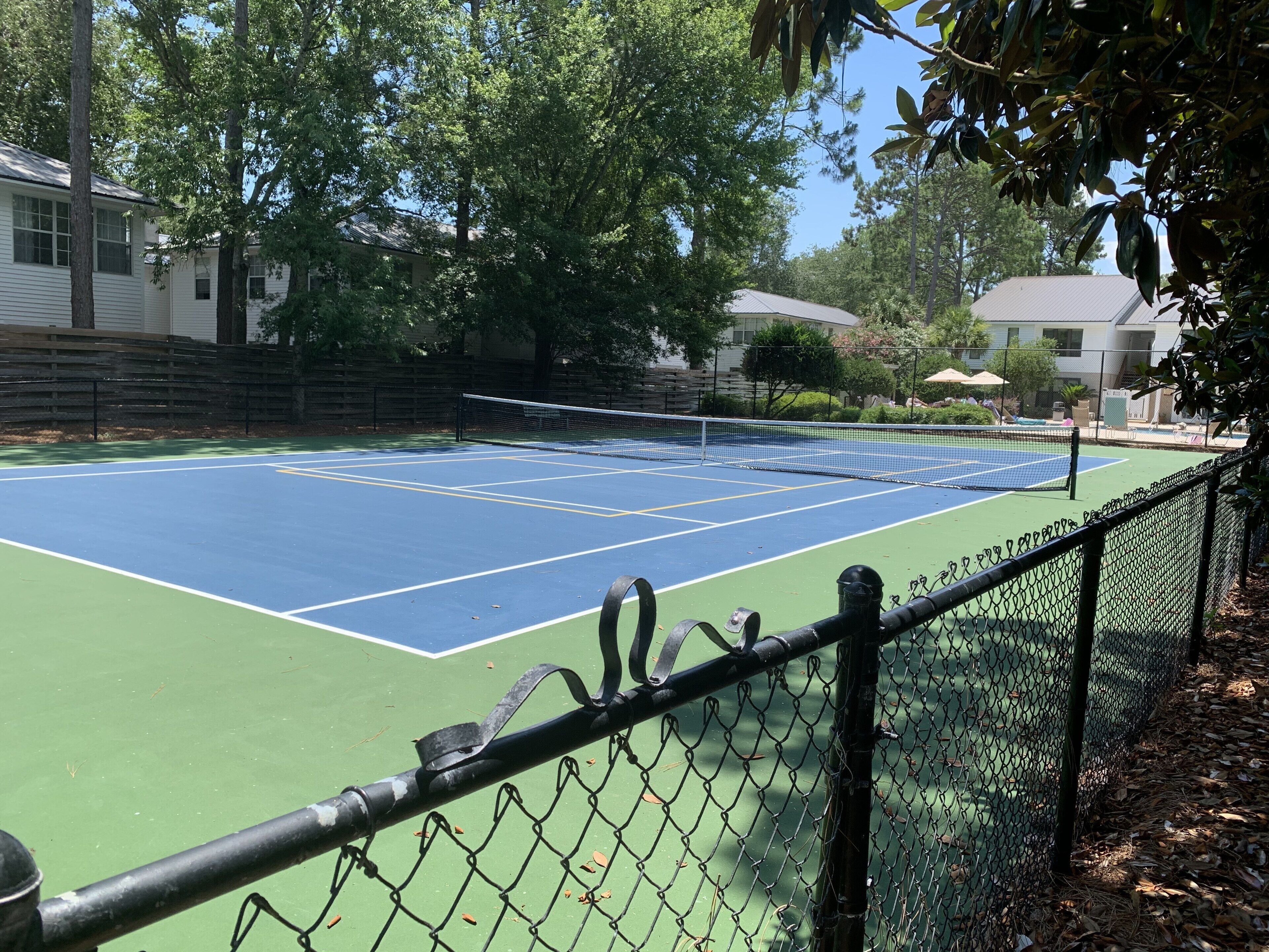 Un court de tennis entouré d’une clôture en treillis métallique, bordé d’arbres et de bâtiments.