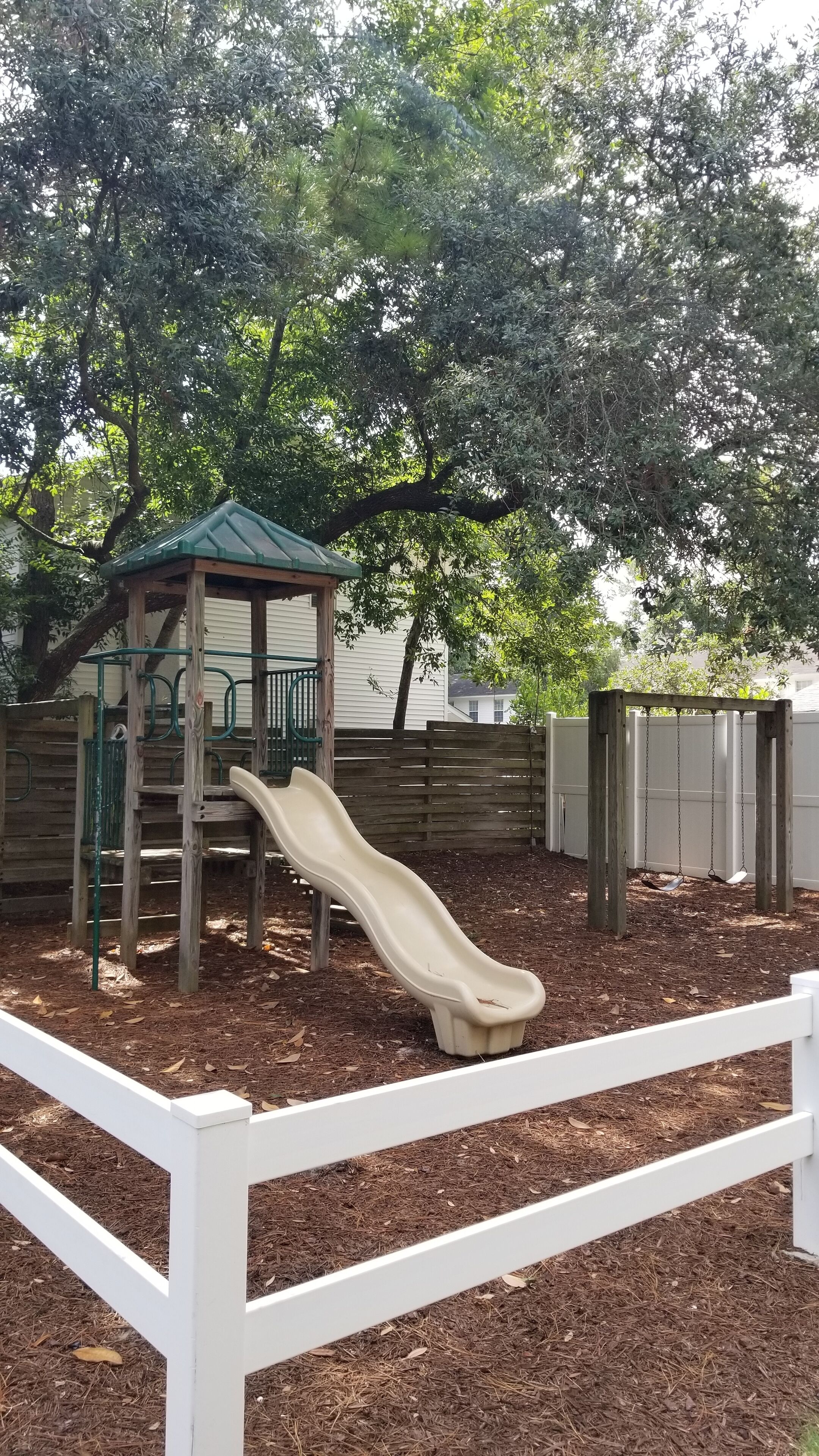 A playground with a slide and climbing structures, surrounded by a white fence and trees.